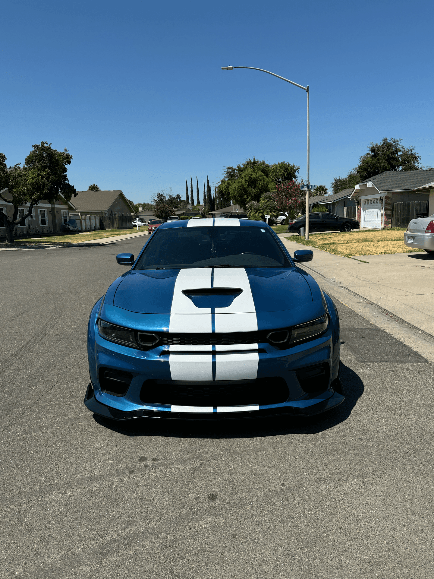 Blue sports car with white racing stripes parked on a residential street on a sunny day.